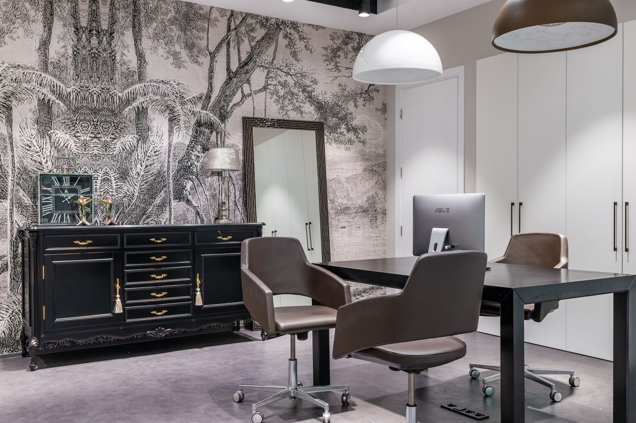 Modern computer on black table near armchairs placed in living room near cabinet with clock in spacious apartment with mirror and closet